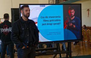Mężczyzna z brodą, ubrany w czarną kurtkę, prowadzi prezentację. Stoi obok ekranu z napisem &bdquo;Czy do stworzenia filmu potrzebny jest drogi sprzęt?&rdquo;.