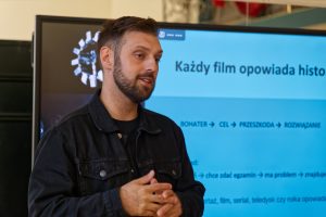Prowadzący tłumaczy zagadnienia, a na ekranie za jego plecami widnieje schemat budowania historii: &bdquo;BOHATER -> CEL -> PRZESZKODA -> ROZWIĄZANIE&rdquo;.