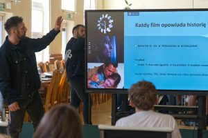 Prowadzący tłumaczy zagadnienia, a na ekranie za jego plecami widnieje schemat budowania historii: &bdquo;BOHATER -> CEL -> PRZESZKODA -> ROZWIĄZANIE&rdquo;.