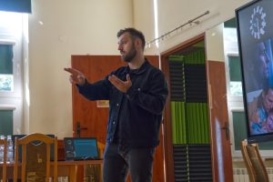 Mężczyzna z brodą, ubrany w czarną kurtkę, prowadzi prezentację.