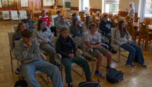 Sala konferencyjna z wieloma młodymi ludźmi siedzącymi na krzesłach w rzędach. Słuchają prezentacji, patrząc w lewą stronę.