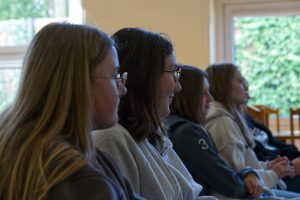 Trzy młode kobiety siedzące w profilu na krzesłach. Patrzą przed siebie, poza kadr, prawdopodobnie słuchając prelekcji.