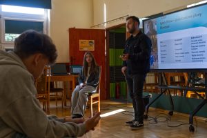 Prelegent stoi przed ekranem z prezentacją o języku filmu i planach filmowych. M&oacute;wi i gestykuluje do uczestnik&oacute;w warsztat&oacute;w. Widoczny jeden siedzący uczeń.