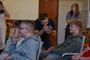 Instruktor warsztat&oacute;w filmowych robi zdjęcie grupie roześmianych uczni&oacute;w siedzących na krzesłach za pomocą smartfona. Obok stoi obserwatorka.