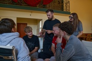 Zbliżenie na instruktora w czarnej koszulce, kt&oacute;ry gestykulując, tłumaczy zagadnienie dw&oacute;m młodym, słuchającym uważnie uczniom.