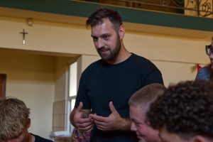 Zbliżenie na instruktora w czarnej koszulce, kt&oacute;ry gestykulując, tłumaczy zagadnienie dw&oacute;m młodym, słuchającym uważnie uczniom.