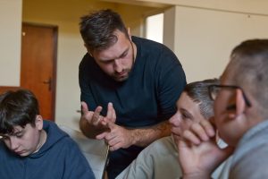 Zbliżenie na instruktora w czarnej koszulce, kt&oacute;ry gestykulując, tłumaczy zagadnienie dw&oacute;m młodym, słuchającym uważnie uczniom.