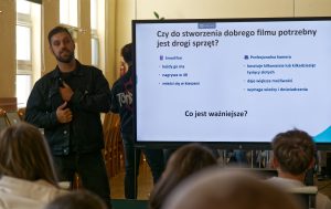 Prelegent trzyma dłoń na klatce piersiowej, stojąc przy ekranie. Slajd prezentuje por&oacute;wnanie sprzętu i zadaje pytanie &bdquo;Co jest ważniejsze?&rdquo;.