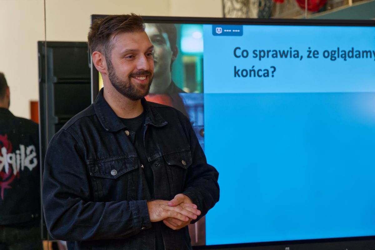 Uśmiechnięty prowadzący w czarnej kurtce stoi obok ekranu wyświetlającego slajd z nagłówkiem „Co sprawia, że oglądamy do końca?”.