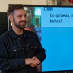 Uśmiechnięty prowadzący w czarnej kurtce stoi obok ekranu wyświetlającego slajd z nagłówkiem „Co sprawia, że oglądamy do końca?”.